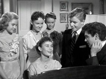 The attendees of the Abernathy's party gather around the piano to admire Laura's (Roberta Shore) playing and singing.