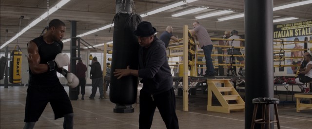 Rocky Balboa (Sylvester Stallone) holds a punching bag for Adonis Creed (Michael B. Jordan) while others look on at the training session.