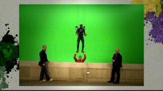 Joined by several crew members, Aaron Johnson in full Kick-Ass gear dangles from wires in a front of later-to-be-added skyline in a photo from the "Taking Flight" gallery.