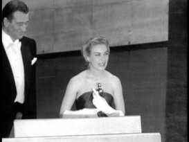 A happy John Wayne looks on as Joanne Woodward accepts the Academy Award for Best Lead Actress in a Fox Movietone newsreel clip.