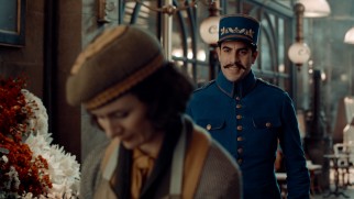 Vigilant inspector Gustave Dasté (Sacha Baron Cohen) practices a smile on station florist Lisette (Emily Mortimer).