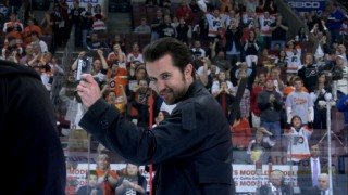 Mac (Rob McElhenney) relishes the chance to look cool while taking his center ice slap shot at a Flyers game.