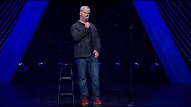 Jim Gaffigan sports black shirts, blue jeans, and red shoes against a black and blue set.