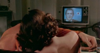 A flustered newscaster (Neil Thompson) watches on from the television as a young couple undresses and grows amorous on the couch before him.