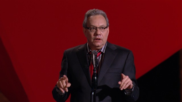 Lewis Black performs in front of a giant suspended red cube in his stand-up comedy special "In God We Rust."