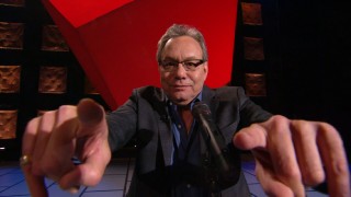 With fingers extended toward the camera, Lewis Black advertises "In God We Rust" as a 3D presentation, which it is not.