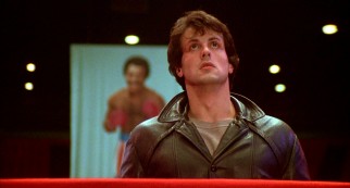 With Apollo Creed smiling behind him, Rocky (Sylvester Stallone) looks up to the rafter at the banner bearing his own likeness.