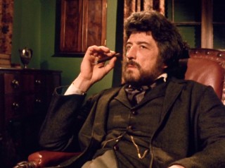 Samson Flusky (John Hallam) listens to conflicting accounts with a thin cigar in his hand.